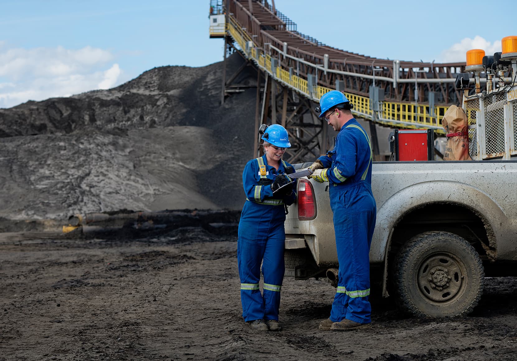 Two workers, Michelle Liebau and Mike Ellis, talking beside a truck by the main feed conveyor from the crusher at Extraction Plant 86. They are wearing blue hard hats, safety glasses, ear plugs, coveralls and gloves.