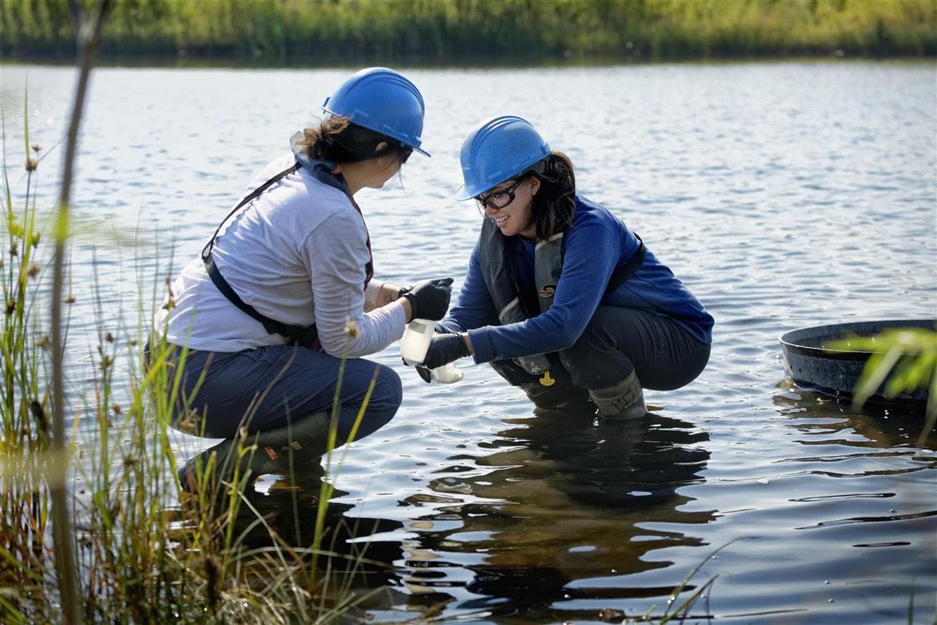 Two women in hard hats crouching in a pond while testing the water
