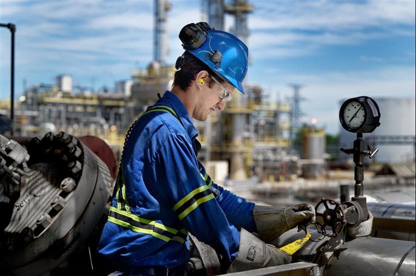Male worker at the Sarnia Refinery. He is wearing a blue hard hat, safety glasses, ear plugs, gloves and coveralls. He is checking a yellow tag on a valve. 