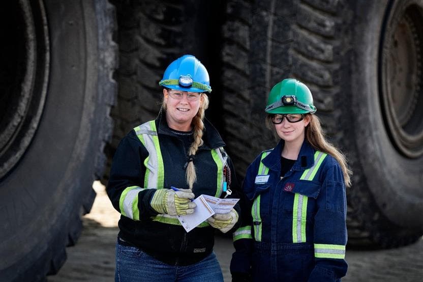 Two female heavy equipment operators wearing protective clothing, hardhats and safety glasses standing in front of two large tires.