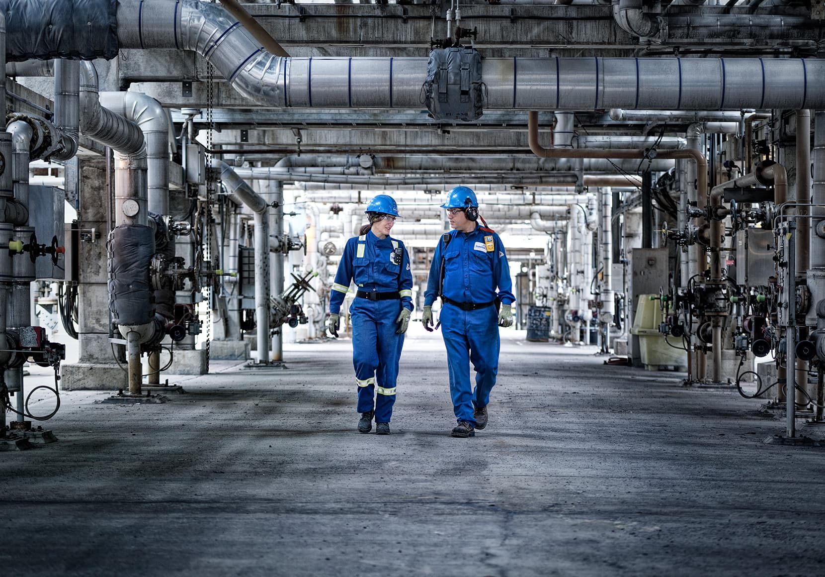 Two workers - Marie-Pier Lauzé and Kalifa Dahani - walking at Unit 1 of the Montreal refinery. They are wearing blue hard hats and gloves. Marie-Pier is also wearing coveralls.