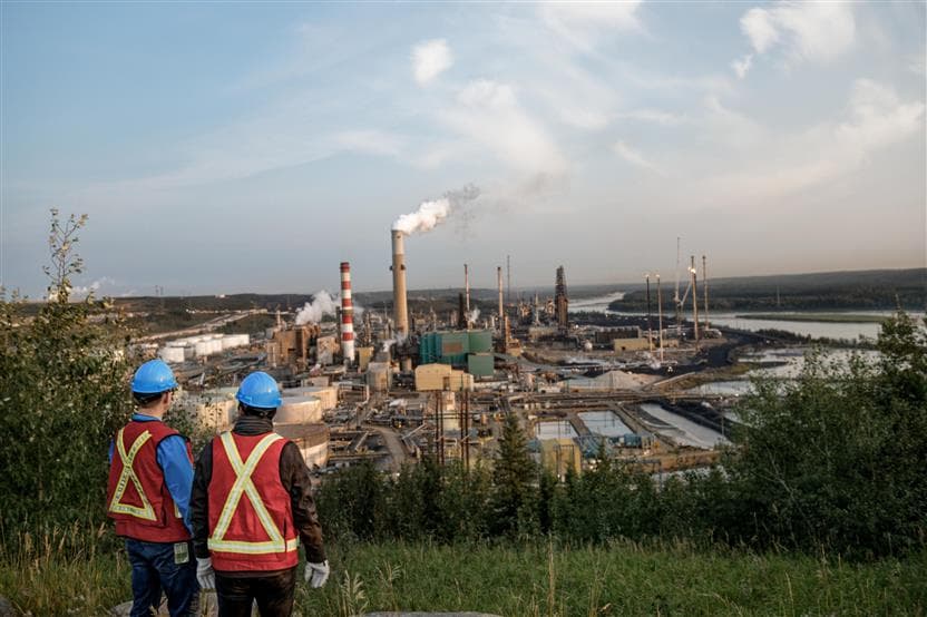 Two male workers standing with their backs to the camera. They are on a hill looking down at Base Plant. Both are wearing blue hard hats, safety vests and gloves.