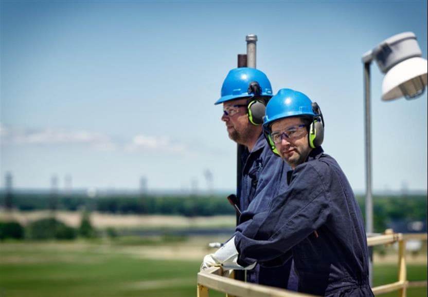 Two male workers standing on top of a silo at the St. Clair Ethanol Plant. They are wearing blue hard hats, safety glasses, coveralls and gloves.