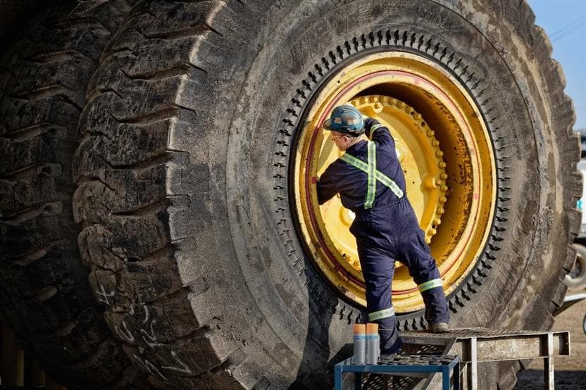 A male worker, Tony Bussey, changing a tire on a haul truck at the Mine Equipment Maintenance truck shop. He's wearing a blue hard hat, coveralls, safety glasses and gloves.