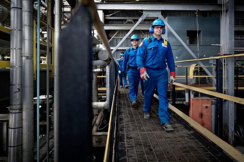 Three workers - Étienne Leduc, Guy Rainville and Lyes Debbon - walking on a walkway at the Montreal refinery. They are wearing blue hard hats, coveralls, safety glasses and gloves.