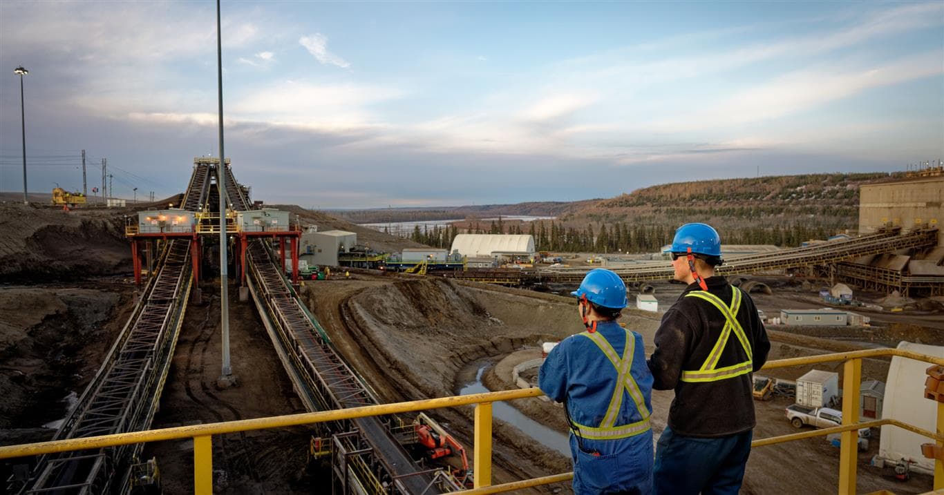 A female and a male worker who are wearing coveralls and PPE are standing at a railing on the top of Plant 82 Extraction facilities looking out towards the river. Below them are the conveyor belts that carry the oil sands to the plant.