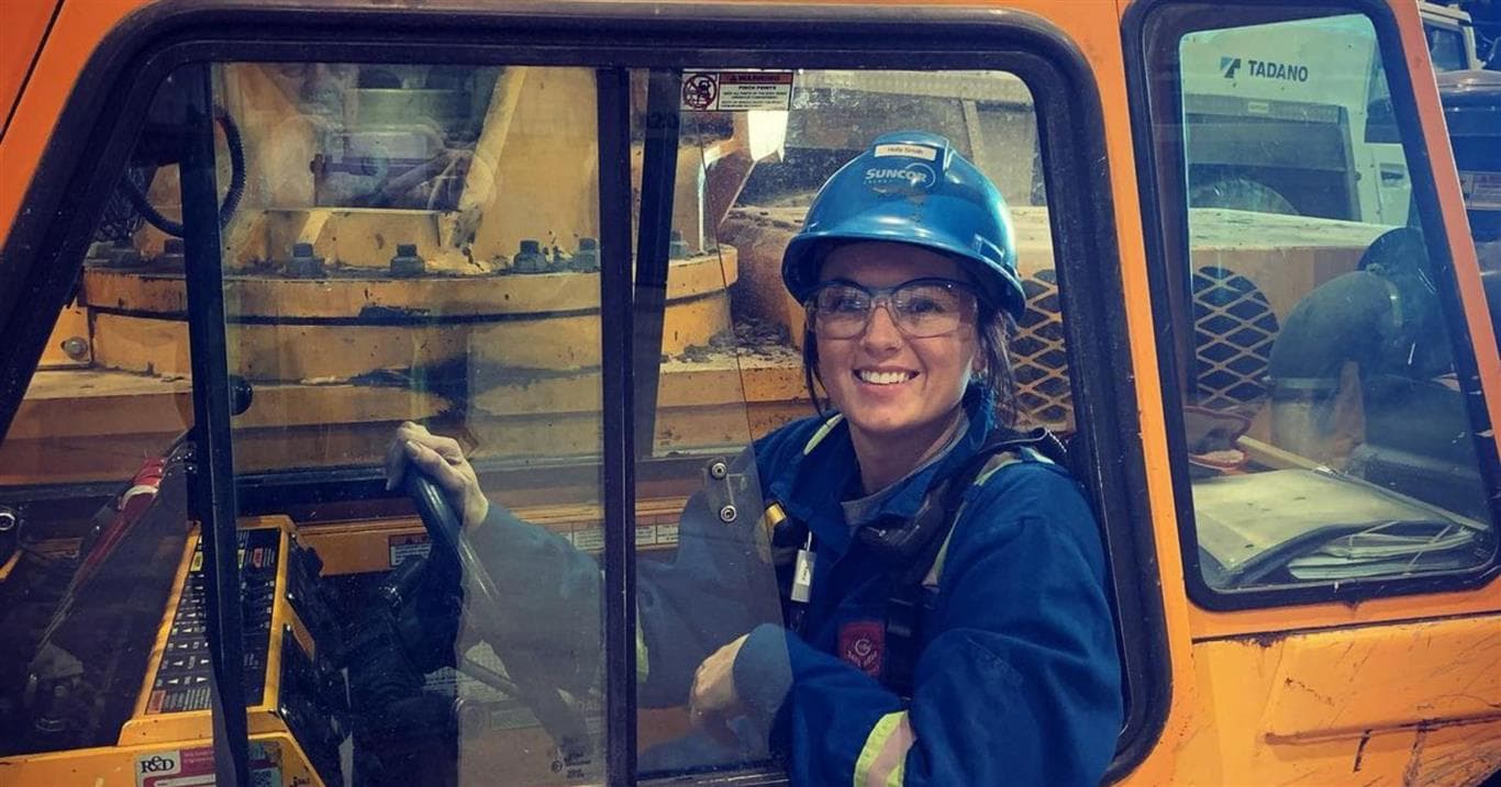 A female crane operator sitting in the operating seat of an 9-ton Broderson Carry Deck crane. The crane is orange in colour, and she’s wearing blue PPE, a blue hard hat and safety glasses.