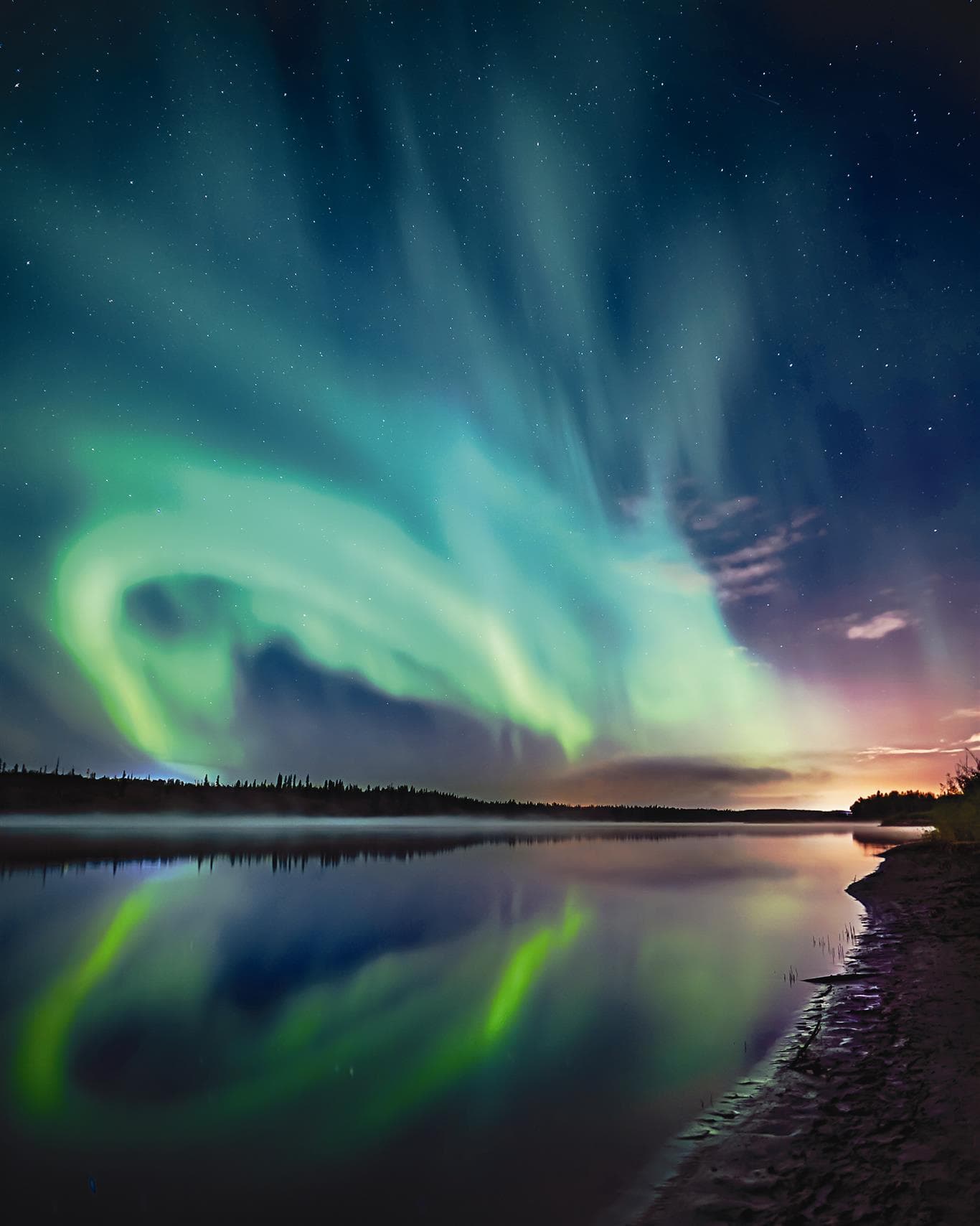 The picture shows the aurora borealis over a river. The sky is dark and clear, and lit with blues and greens.