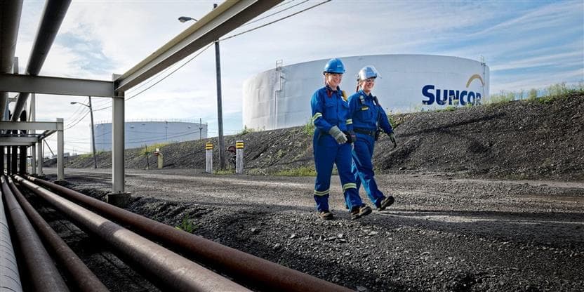Two people in blue coveralls walking outside near pipelines. There is a large, white tank with the Suncor logo in the background and a blue sky.