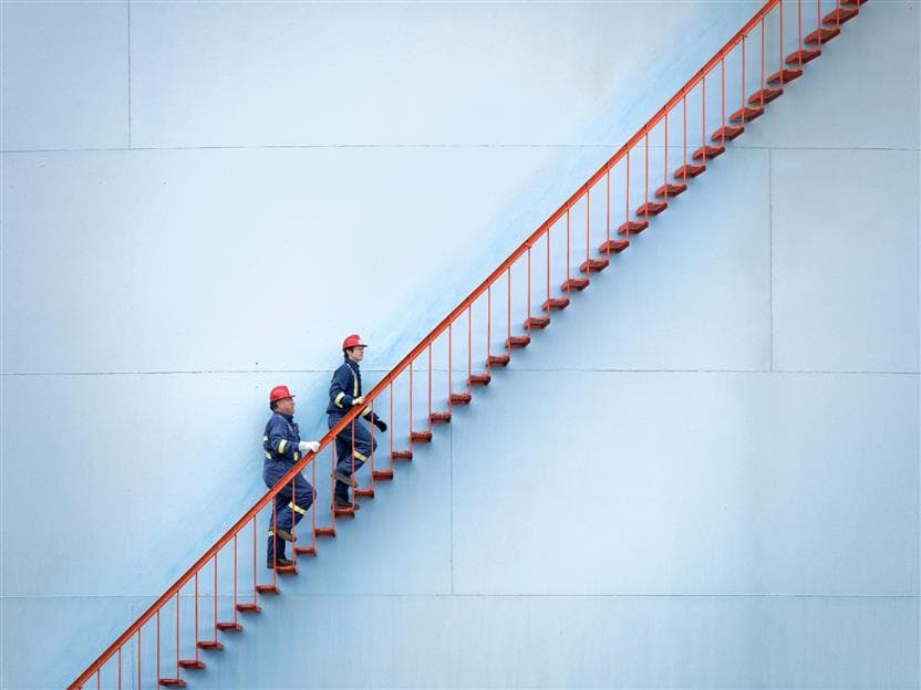 Two male workers, climbing up the red stairs of a holding tank at the Edmonton refinery. They are wearing blue PPE, red hard hats safety glasses and gloves.