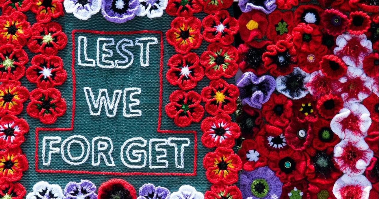 A blanket of crocheted mostly red poppy flowers surround the words, "lest we forget."