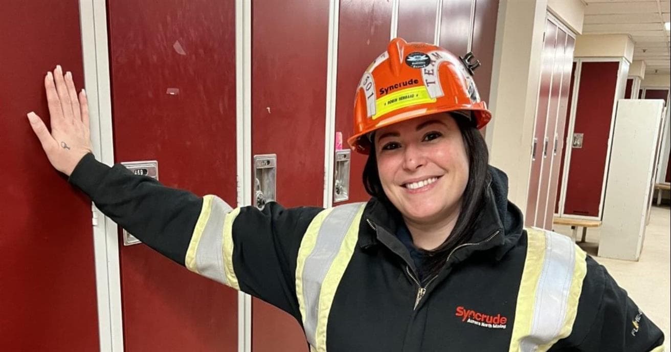 A female wearing a hard hat and a black Syncrude jacket with reflective stripes smiles as she stands in a locker room. She has one hand on her him and the other is pressed against the red lockers.