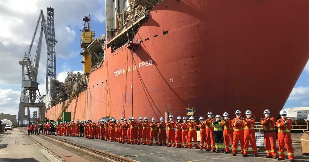 A row of people wearing red coveralls and hardhats stand on a dock. There is a large ship behind them. 
