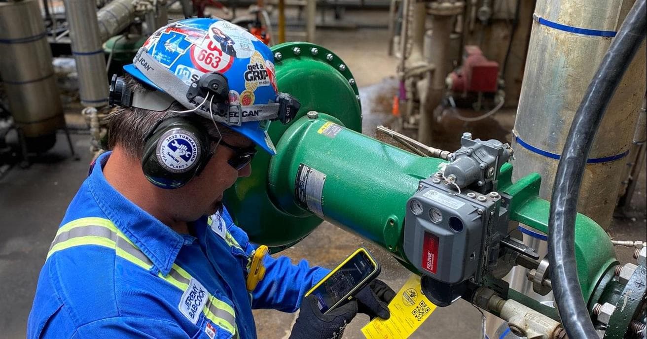 A person wearing a blue shirt with reflective stripes, a hard hat and ear protection uses a smart phone to scan a code on a piece of equipment. 
