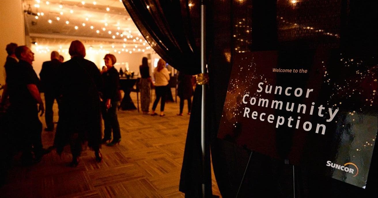 A sign reading “Suncor Community Reception” outside of a ballroom. There are people mingling and small lights hanging from the ceiling. 