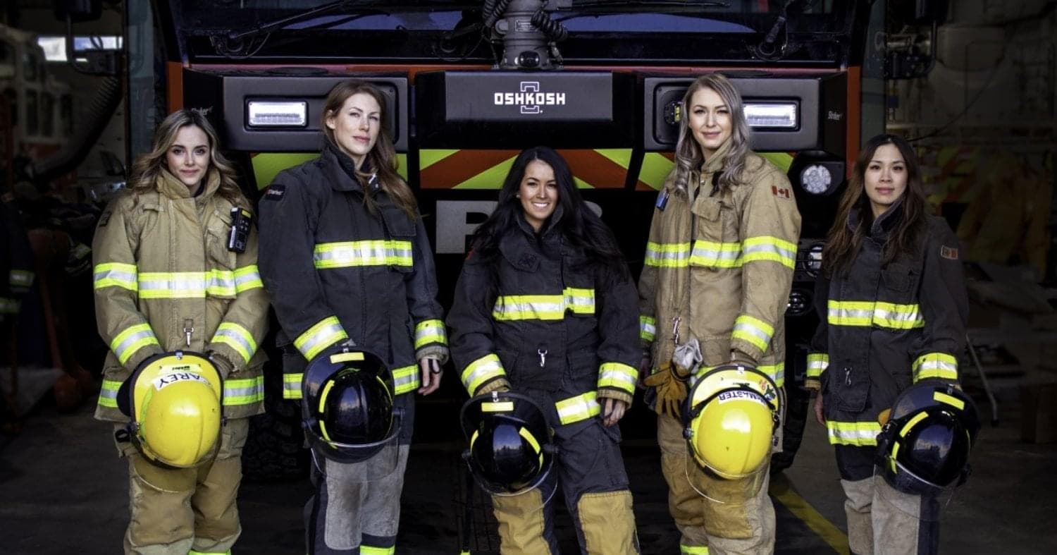 Five women, wearing full-firefighter gear and holding their yellow and black firefighter hats, stand in front of a fire truck. 