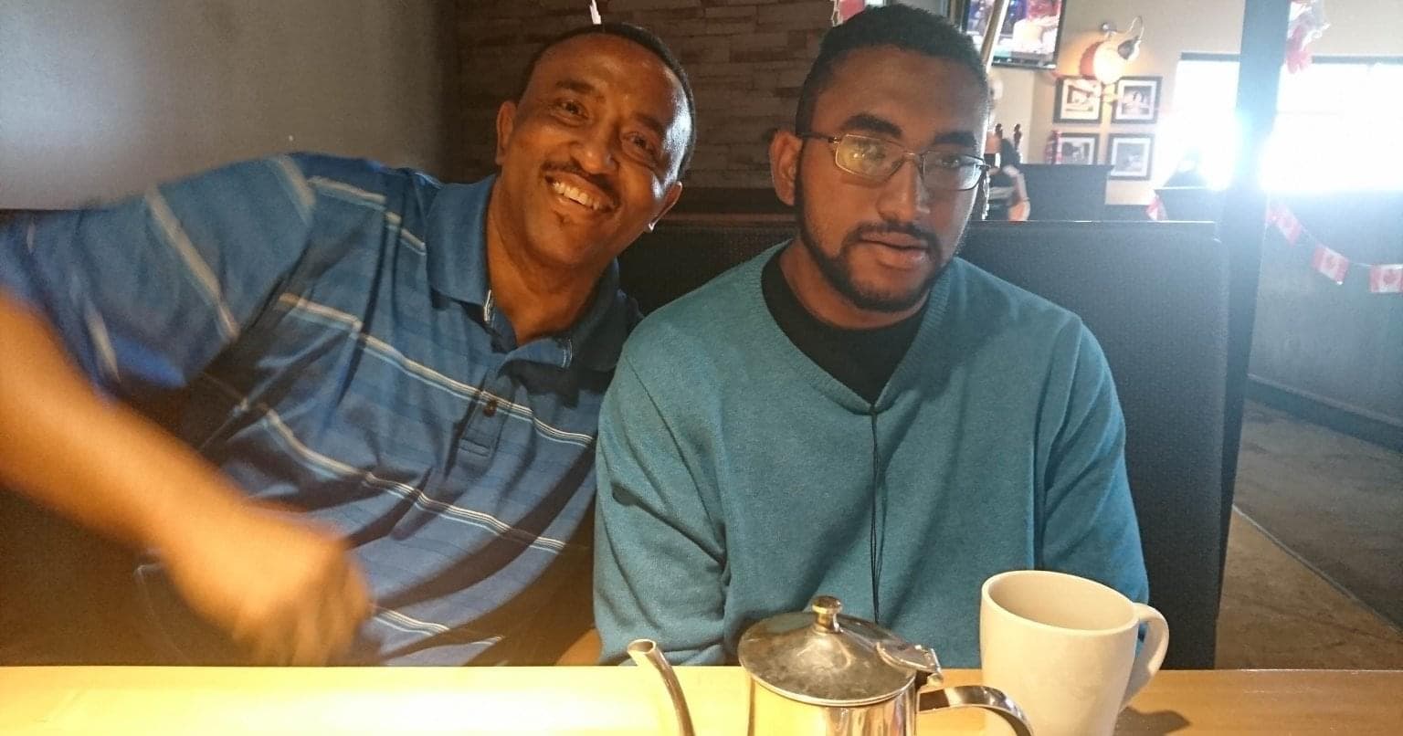  Deux hommes noirs assis sur une banquette dans un restaurant. Les deux portent un chandail bleu. Une théière et une tasse de café se trouvent sur la table. 