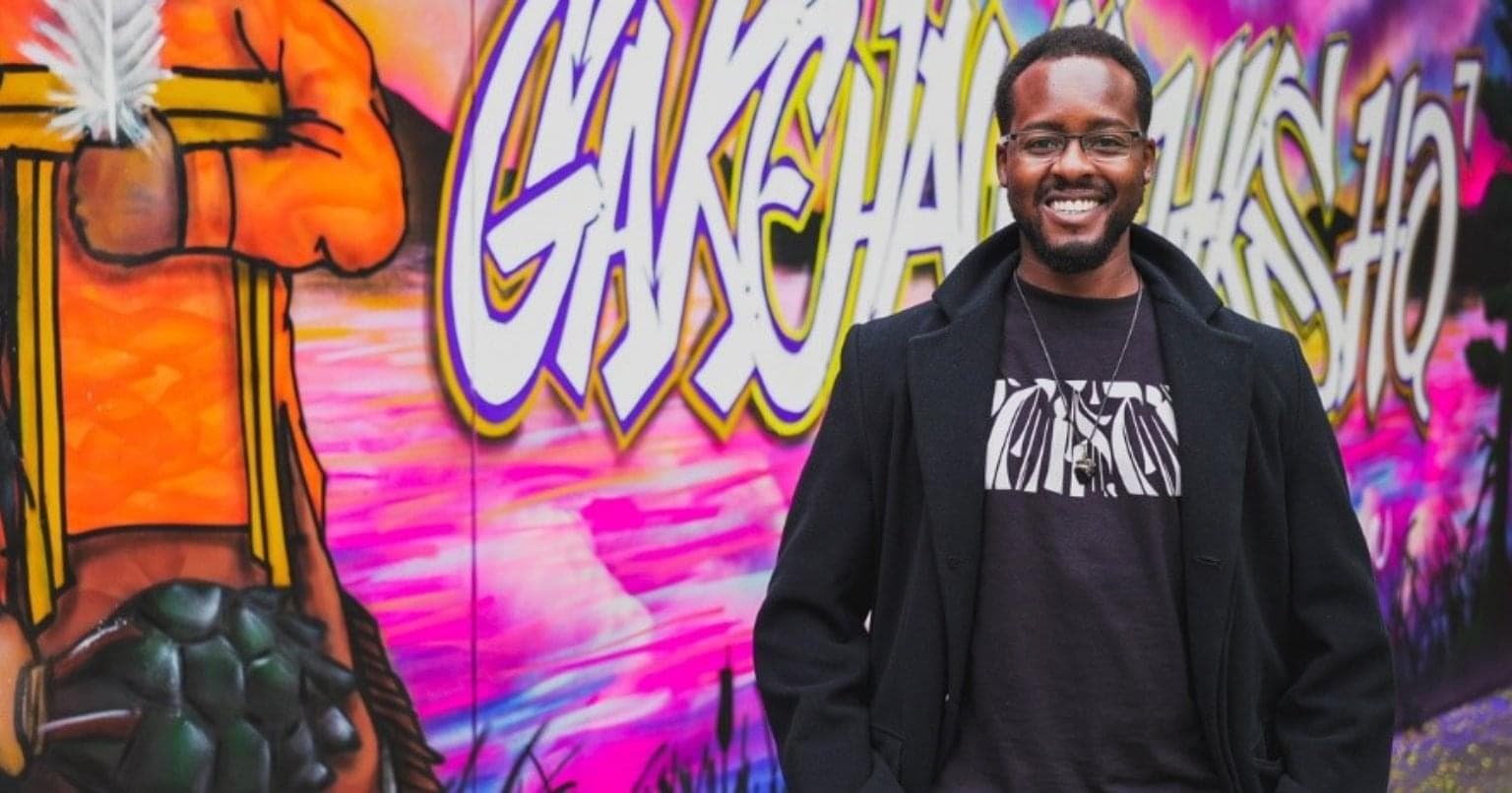 Black Toronto Artist Jessey Pacho standing in front of the colourful mural wearing a black jacket and black shirt.