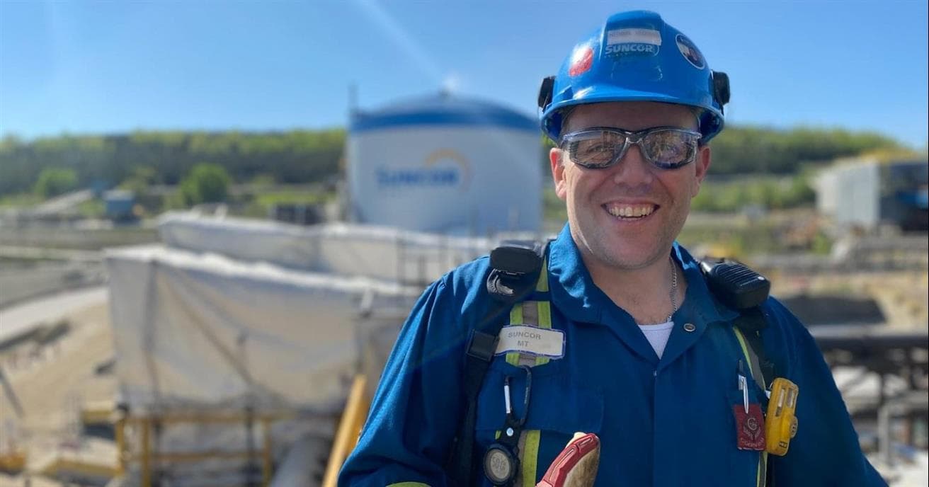 A man wearing blue coveralls, a blue hardhat, safety glasses and gloves smiles at the camera. Behind him are pipes, a large tank, a green hill and blue sky.
