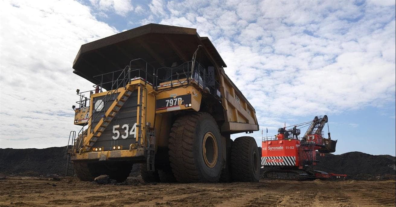 A large, yellow 797 haul truck in an oil sands mine. There is a red shovel behind it and a cloudy sky above.