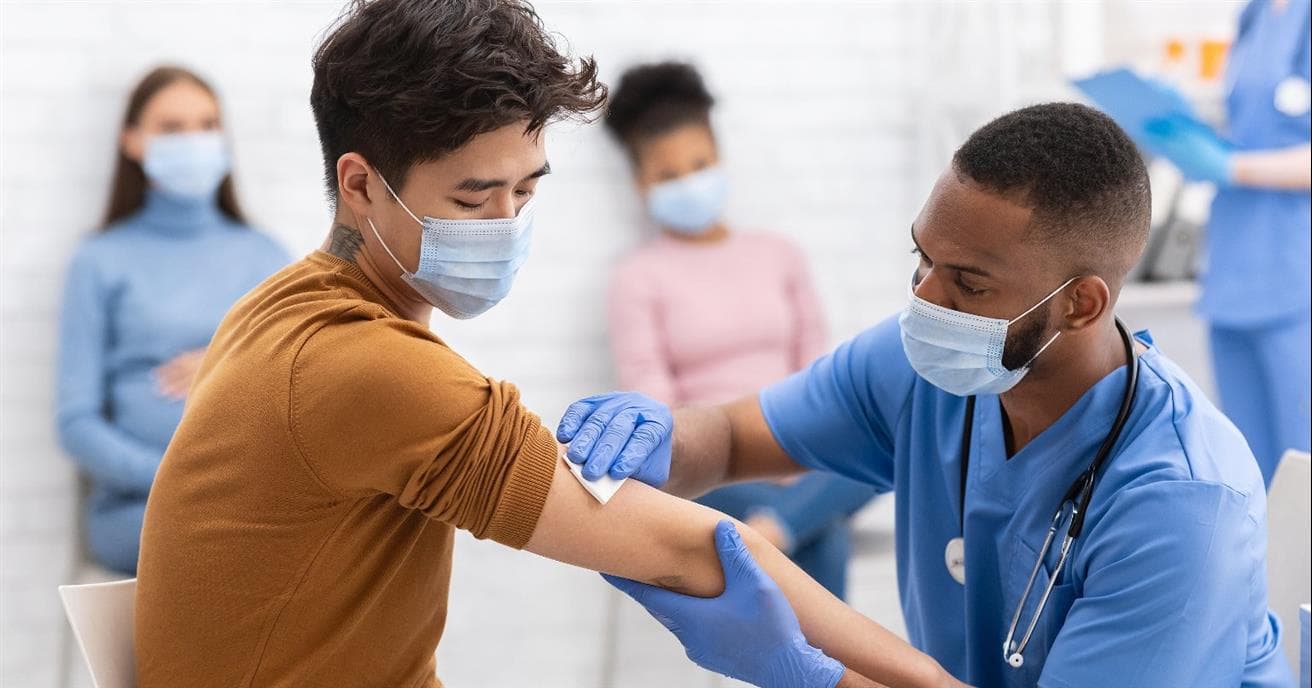 asian man in yellow shirt with his arm stretched out as a black male nurse puts a cloth on it after vaccination