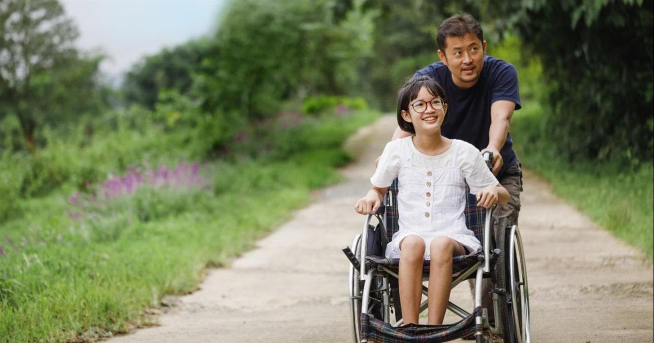 An adult pushing a child in a wheelchair on a path. There is green grass and trees lining the path.