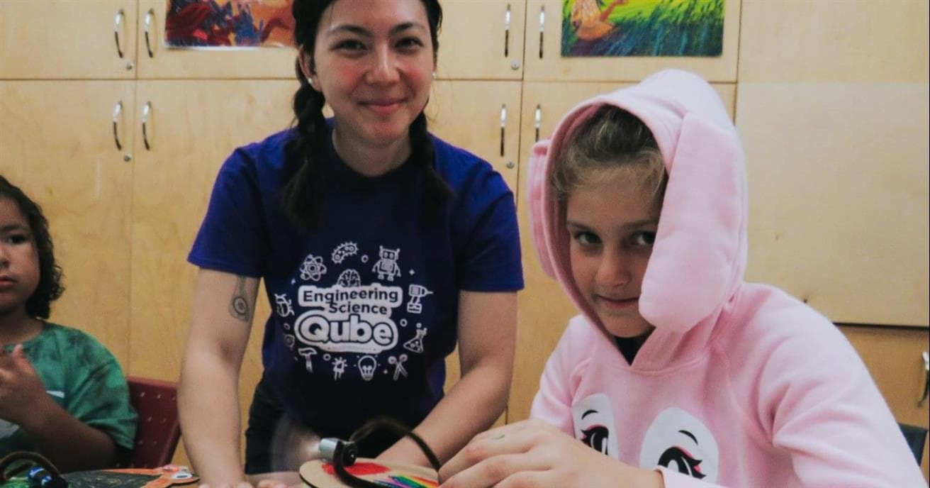 Two people, one child one young adult, smile at the camera while constructing a small robot. One person is wearing a pink sweatshirt with a hood, while the other is wearing a blue t-shirt with “engineering science qube” written on the front. 