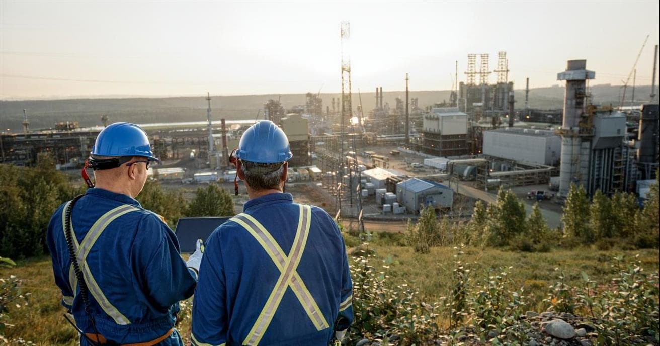 Two men wearing blue coveralls and hard hats overlook an oil sands mine site.