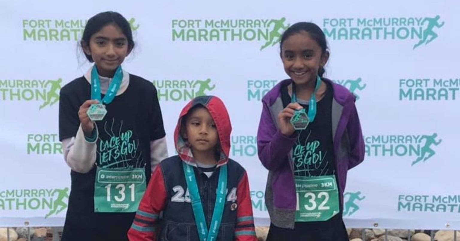 trois enfants souriants avec des médailles autour du cou debout devant un panneau indiquant le marathon de Fort McMurray
