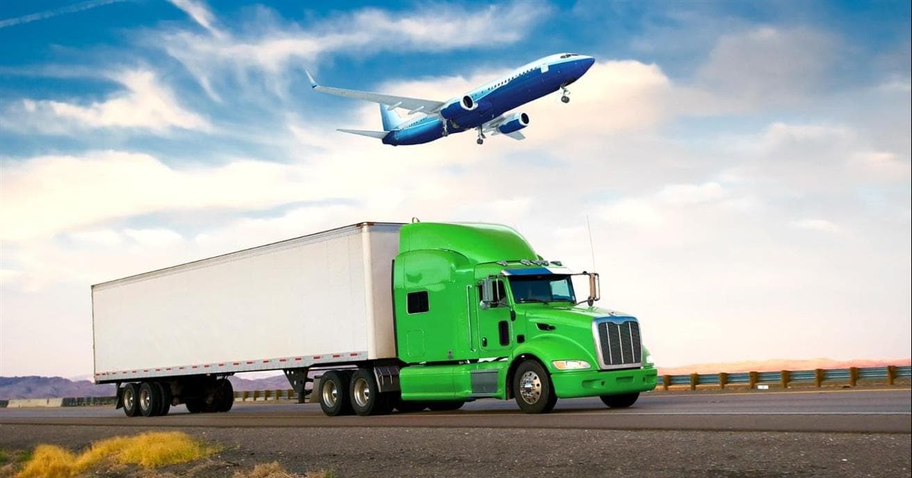 A white and green truck drives along a road with a plane flying in the sky overhead.