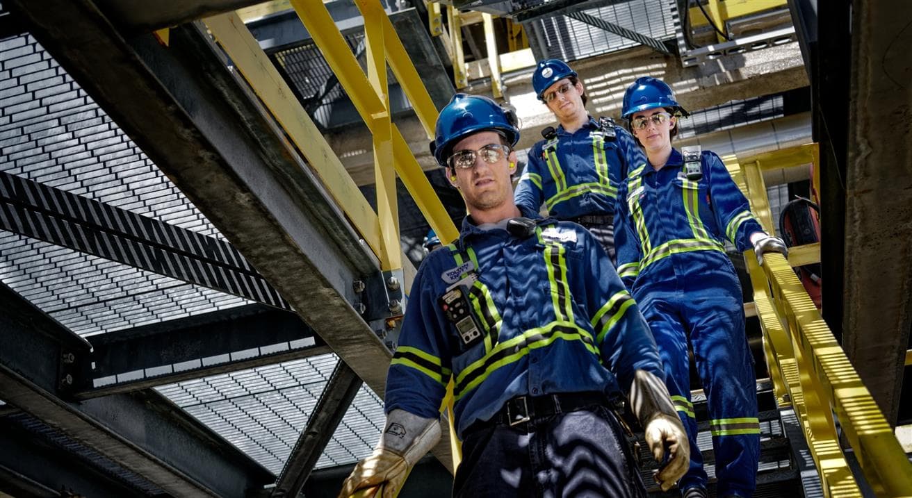 Three people in blue coveralls and hardhats are walking down a set of stairs.