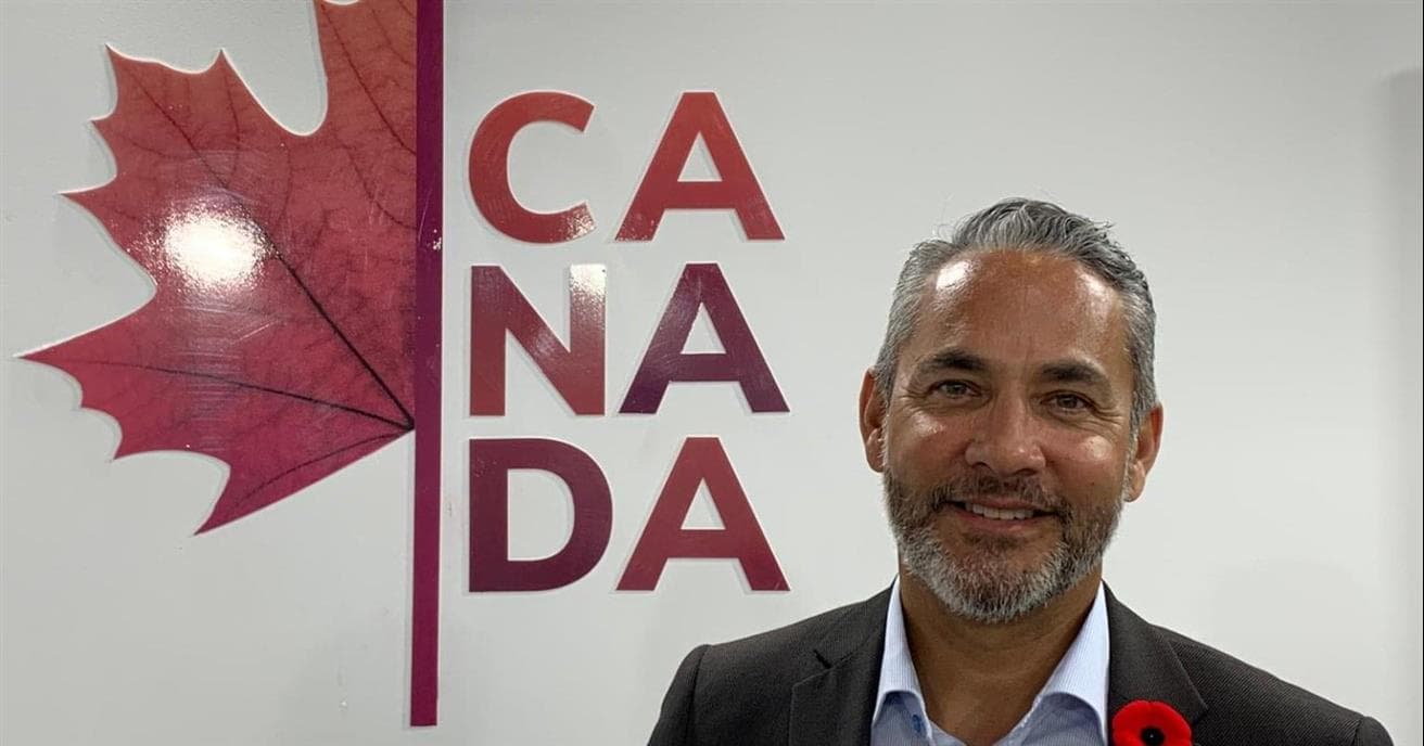 JP Gladu se tient à côté du logo du Canada, qui consiste en une moitié de feuille d'érable avec le mot CANADA de l'autre côté. Il porte un veston et sourit à la caméra.