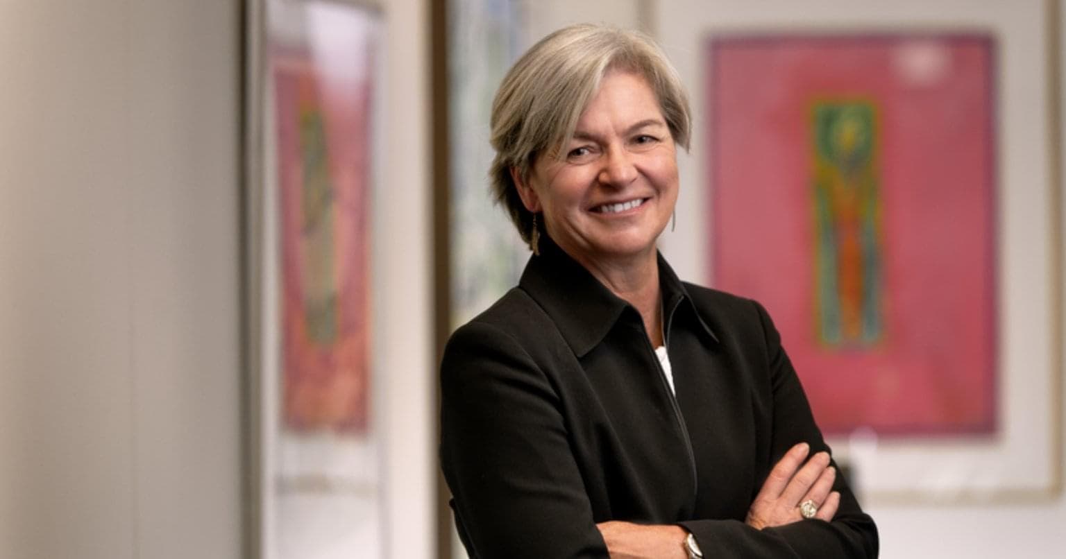 A headshot of Chief Climate Officer Martha Hall Findlay, wearing a black blazer. She is standing in a office hallway with her arms crossed and smiling at the camera. There is artwork behind her. 