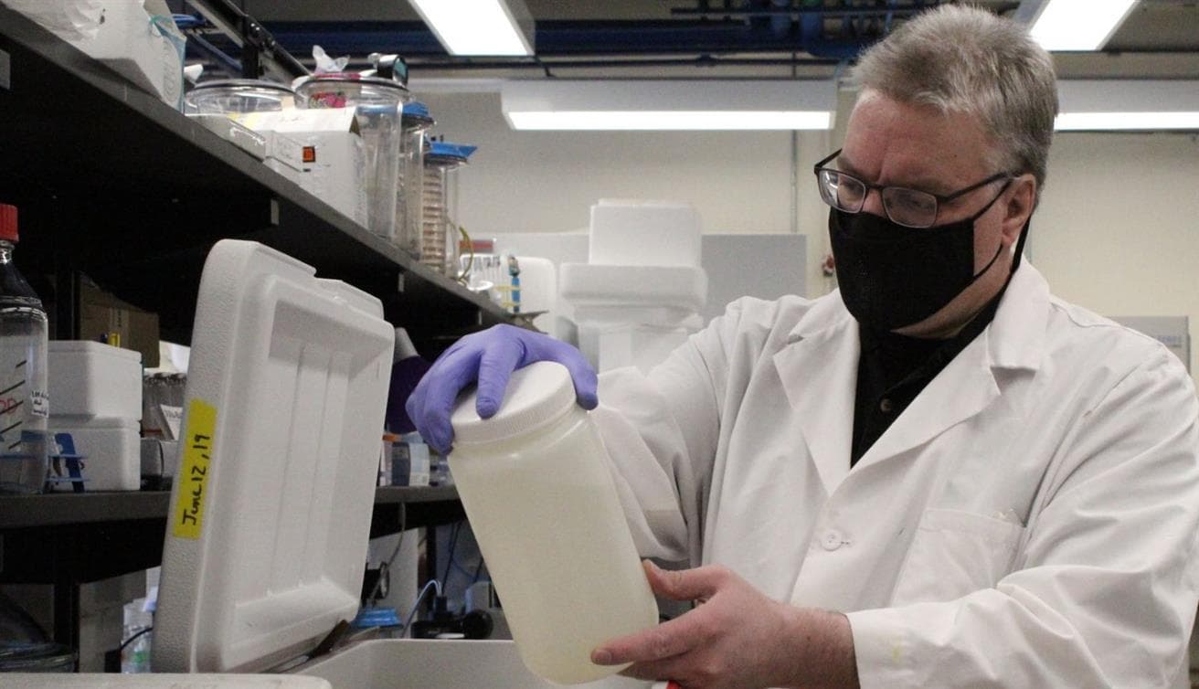 Un homme portant une blouse de laboratoire blanche inspecte un pot qu'il retire d'un réfrigérateur rouge. Il se trouve dans un laboratoire, avec des béchers et d'autres équipements de laboratoire en arrière-plan.