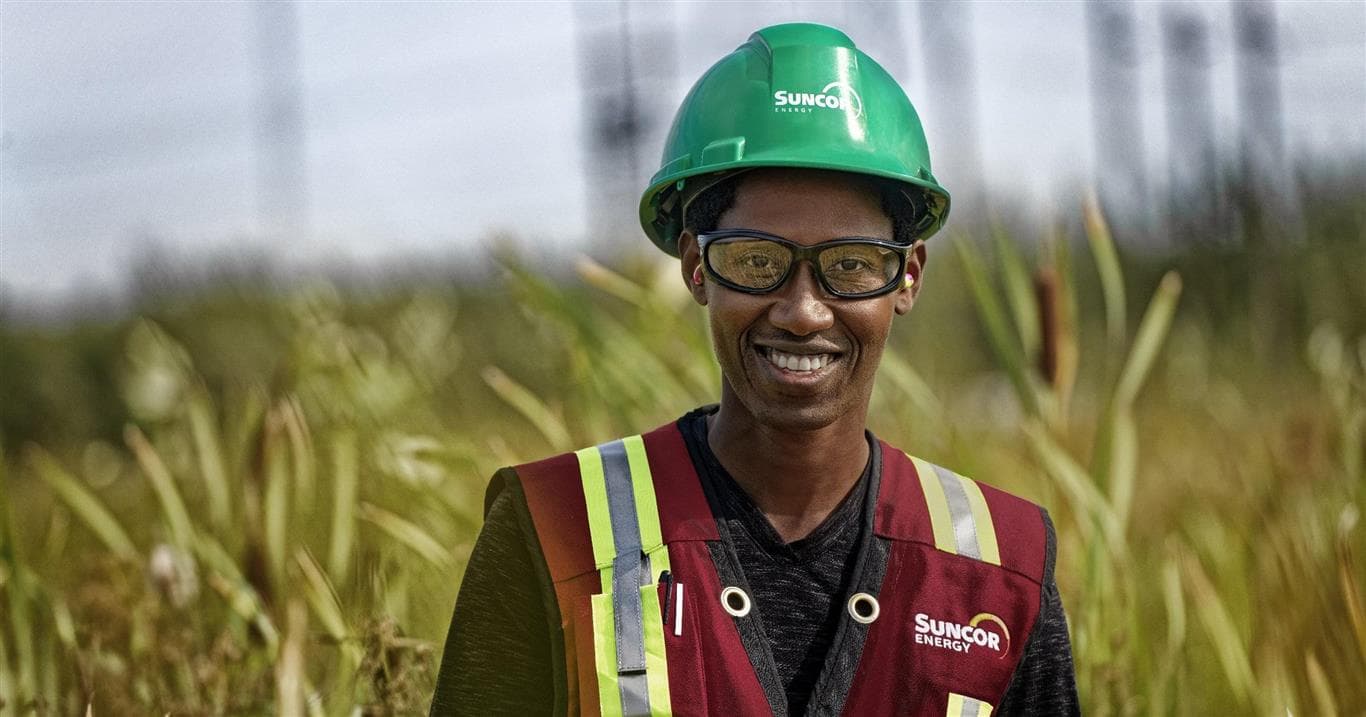 A man wearing a green hardhat and red safety vest standing in a field with tall grass.