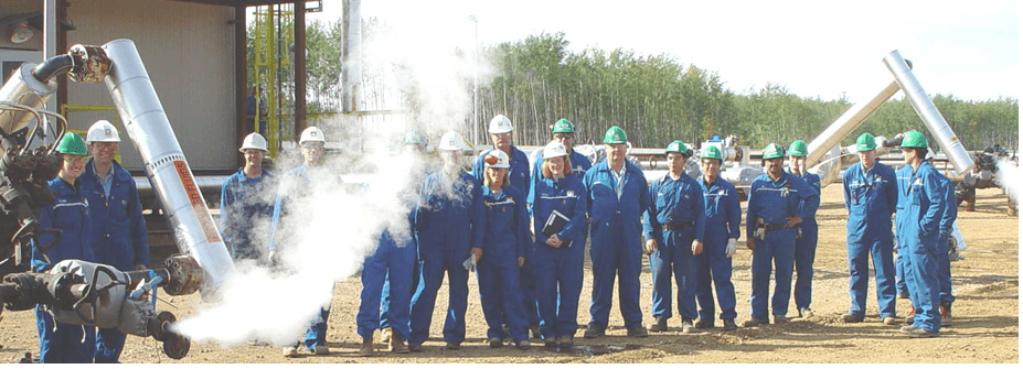 A group of people all wearing blue coveralls and white hard hats. They are outside on a sunny day. There trees in the background and steel pipes emitting steam. 