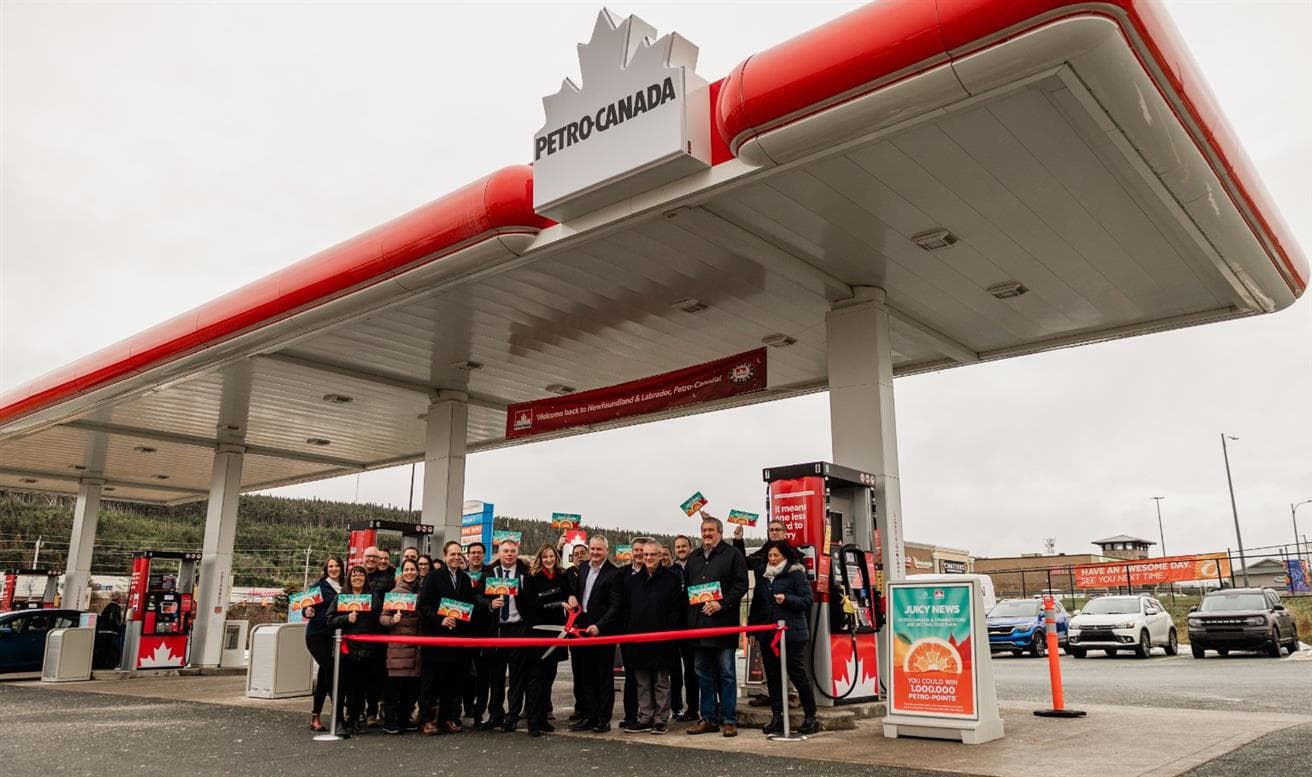 People stand at Petro-Canada fuel pumps with a red ribbon. Someone is cutting the ribbon with large scissors. 
