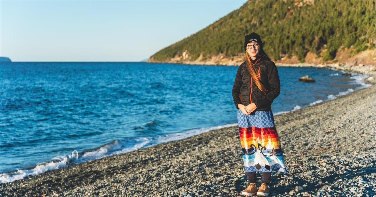 Kelly Young stands by the ocean. She is wearing a colourful, long ribbon skirt and a black jacket.