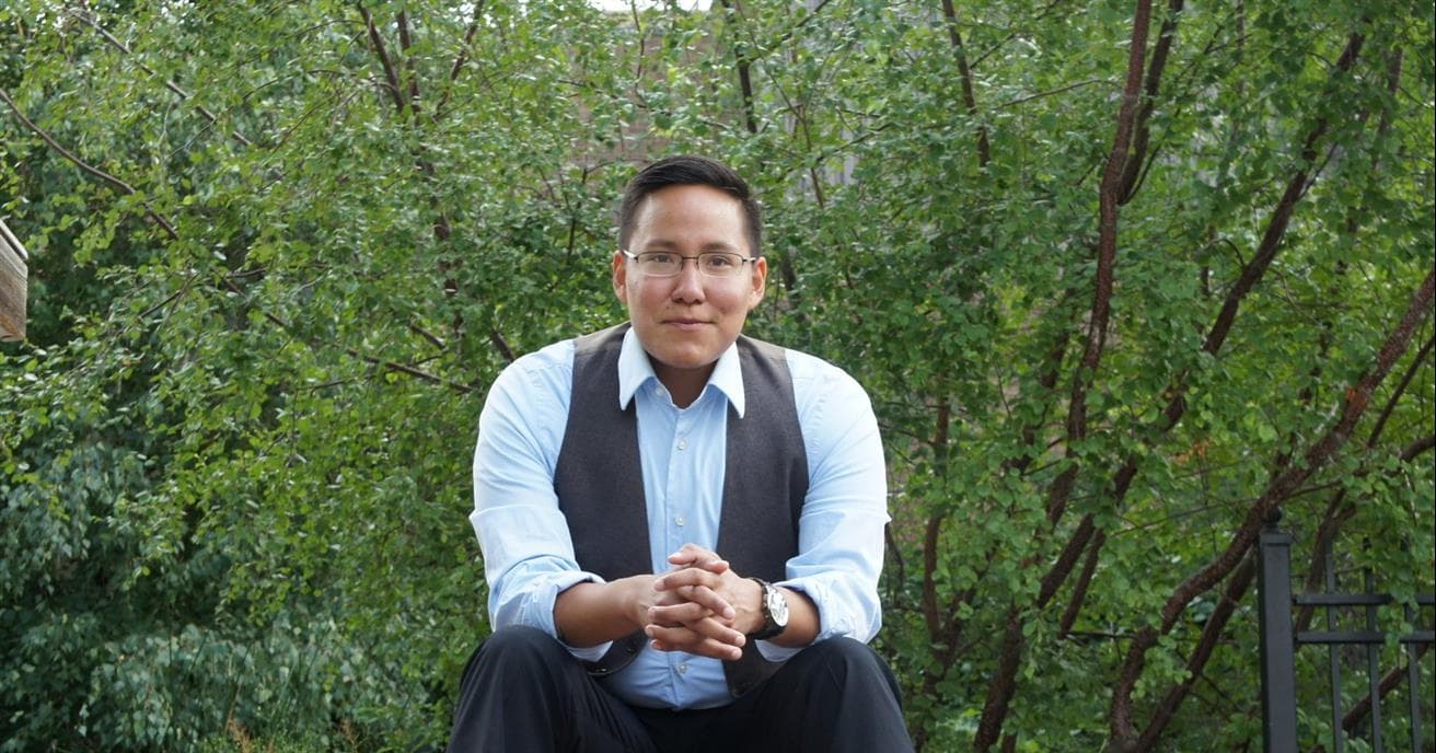 In the photo, a smiling Indigenous man sitting on stone steps wearing a button down shirt with his hands clasped on his knees. 