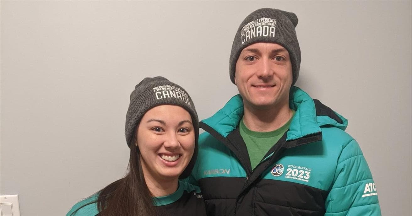 Two people wearing 2023 Arctic Winter Games apparel in black and turquoise, stand together. They are both wearing grey toques and smiling. 