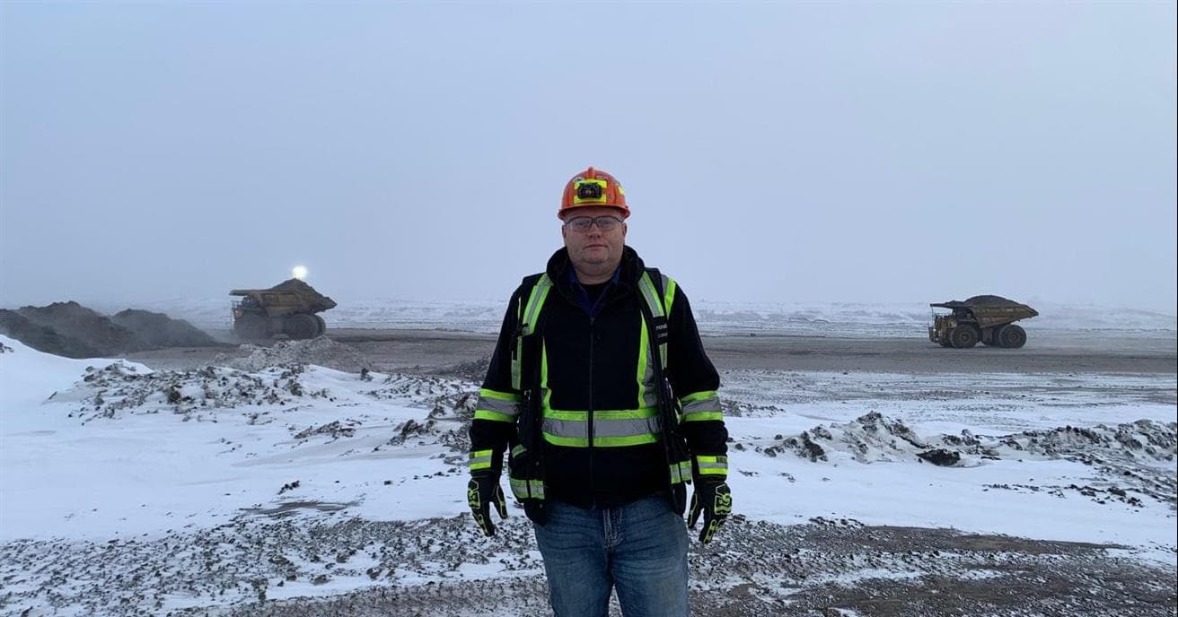 A man wearing a hardhat, safety glasses and winter clothing stands in the foreground in a mine covered with snow with two large haul trucks in the background.