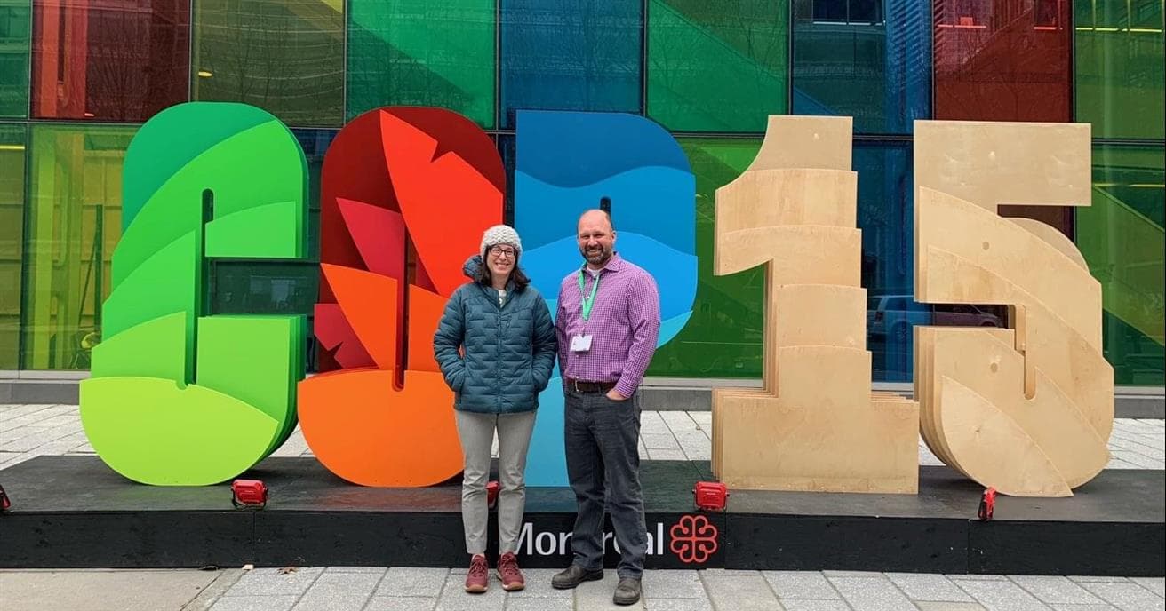 A man and a woman stand in front of a large and colourful COP15 sign. They are outside. The woman is wearing a green puffy jacket and a beige toque, and the man is wearing a purple plaid shirt and dark pants.