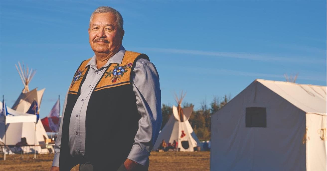 A man stands facing the camera, with a pleasant smile. He is outside in a field; there are tipis and a large white tent behind him. He is wearing a vest with traditional Indigenous beadwork on the top. 