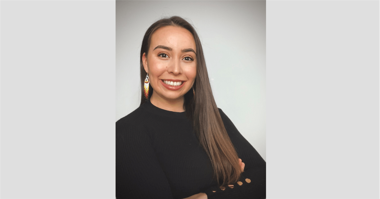A portrait of a young Indigenous woman. The woman is wearing a black sweater and beaded earrings. She is smiling brightly with her arms crossed. 