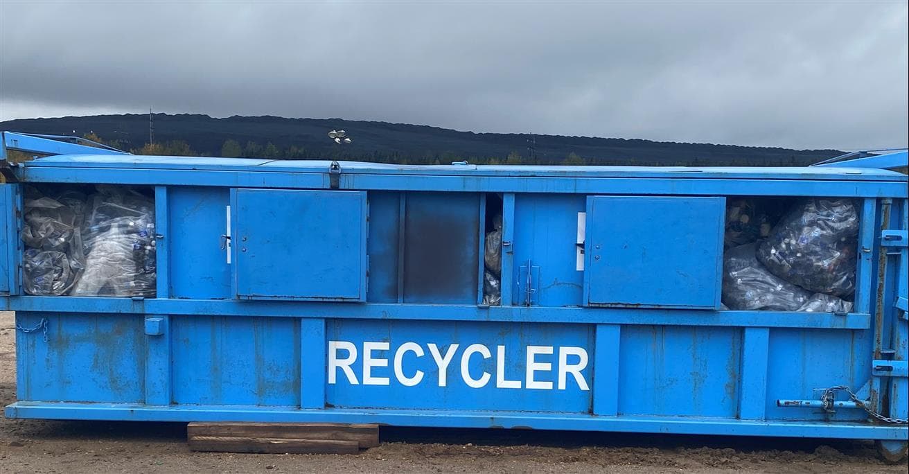 The image is of a very large blue, metal bin with the word “recycler.” The bin is holding many clear recycling bags of empty water bottles 