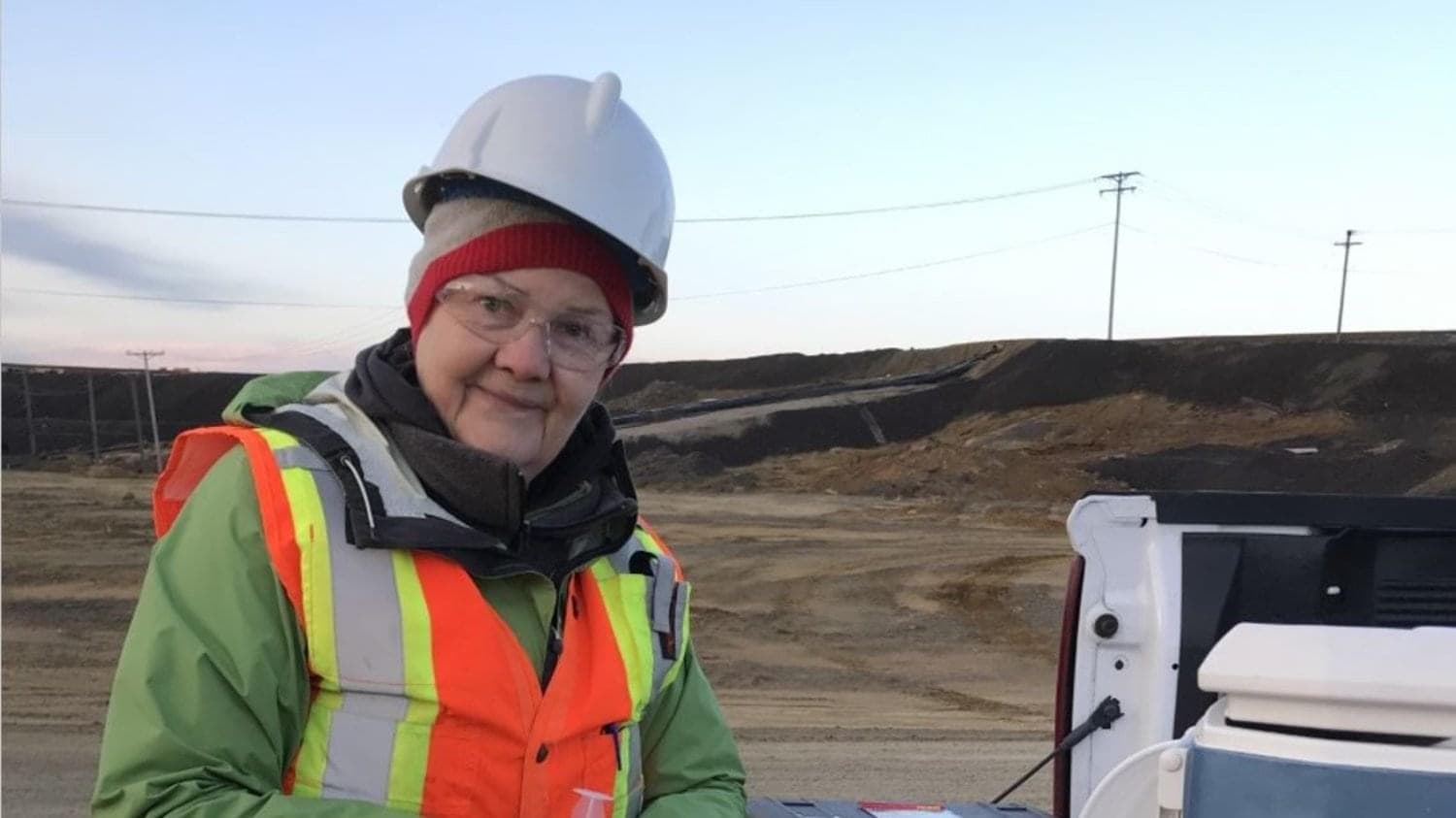 A woman standing at the back of a white truck with its tailgate down. She is wearing a hard hat, a reflective vest, glasses and looking directly at the camera.