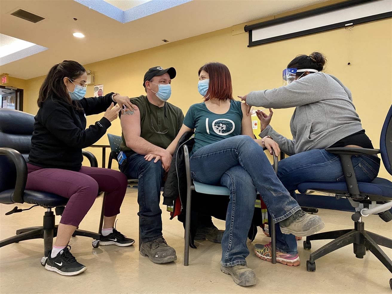 Deux personnes masquées se tenant la main assises pendant qu'elles se font vacciner toutes les deux par des infirmières