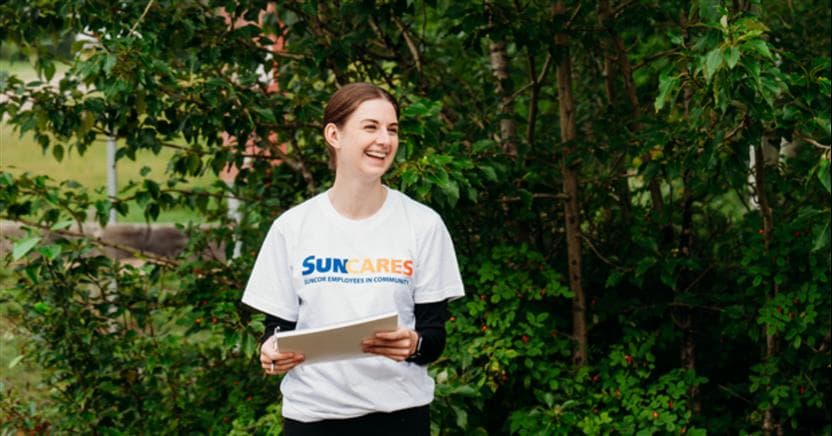A woman is smiling. She is holding a notebook and pen and is wearing a white Suncares volunteer shirt. There are green trees in the background. 