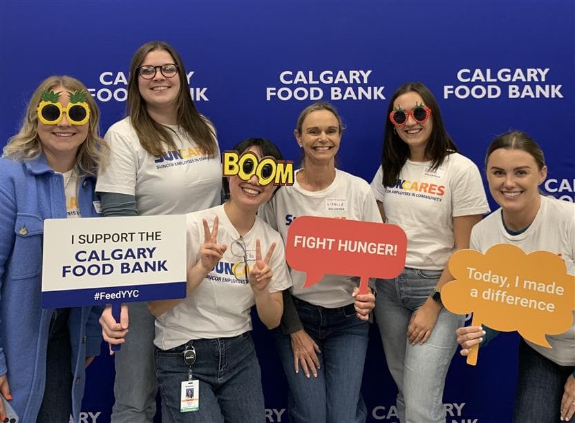 Six women wearing SunCares shirts volunteering at the Calgary Food Bank. They are standing in front of a blue backdrop, and are holding up signs saying “fight hunger” and “today I made a difference”