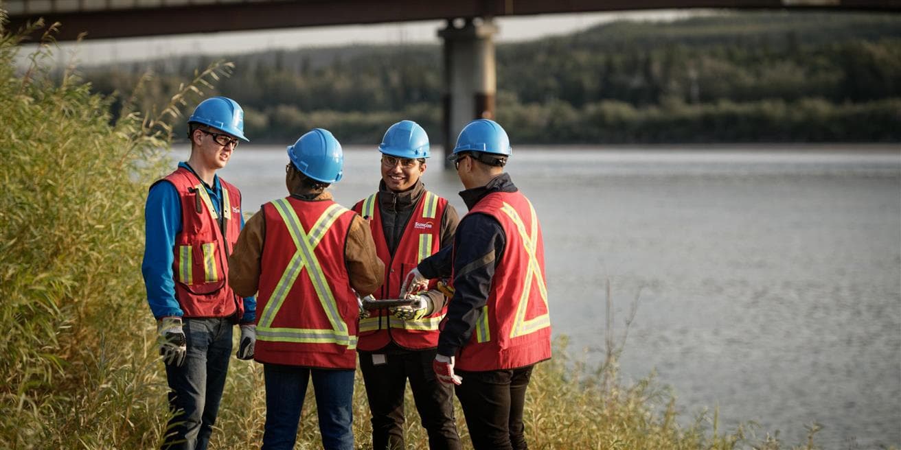 Four researchers standing by the river at Wapisiw lookout. They are wearing safety glasses, blue hard hats, gloves and safety vests. There is a bridge over the river in the background. One of them is holding a tablet. 
